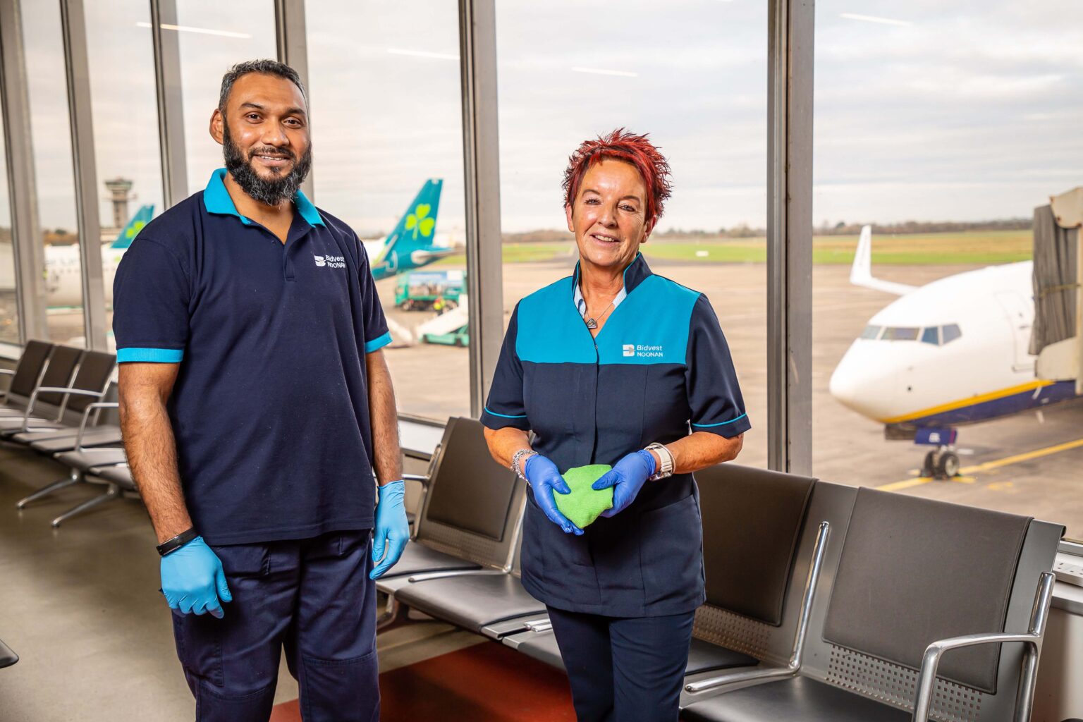 Two airport cleaning staff members stand inside a departure lounge, smiling at the camera. They wear navy and turquoise uniforms and blue gloves. One holds a green cleaning cloth. Behind them, large windows show airplanes parked on the runway.