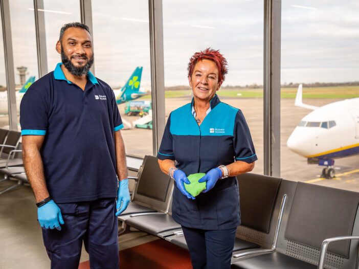 Two airport cleaning staff members stand inside a departure lounge, smiling at the camera. They wear navy and turquoise uniforms and blue gloves. One holds a green cleaning cloth. Behind them, large windows show airplanes parked on the runway.