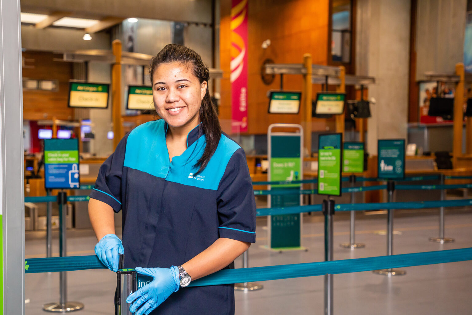 A smiling cleaning staff member wearing a navy and turquoise uniform and blue gloves stands in an airport check-in area, holding onto a barrier post. Check-in signs and queue barriers are visible in the background.